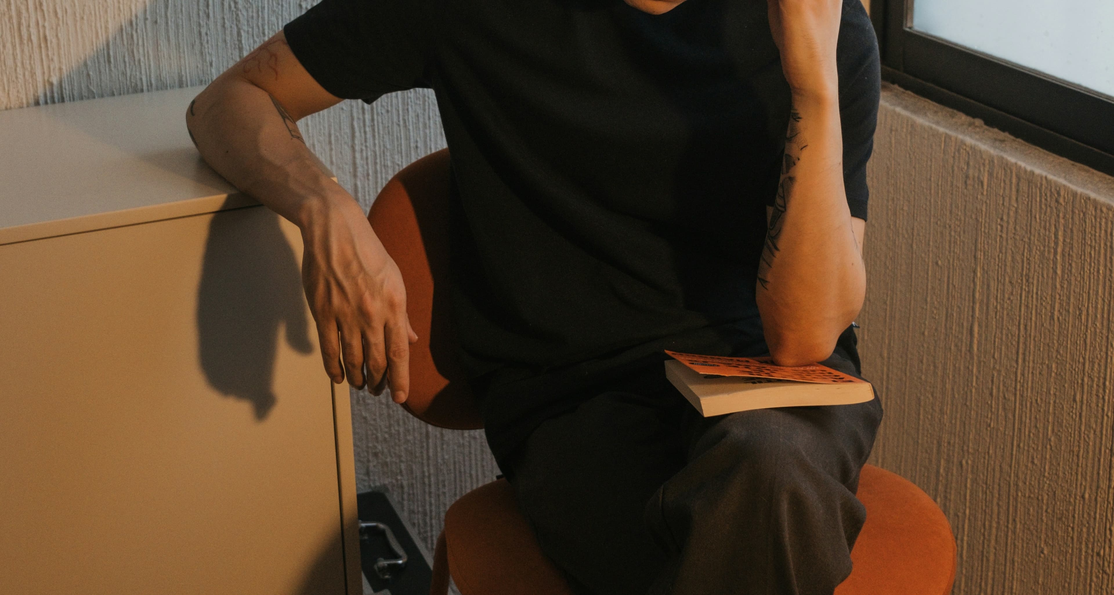 Person in a black shirt sitting on a chair, holding a book, with one hand on their face and a tattoo visible on their arm.