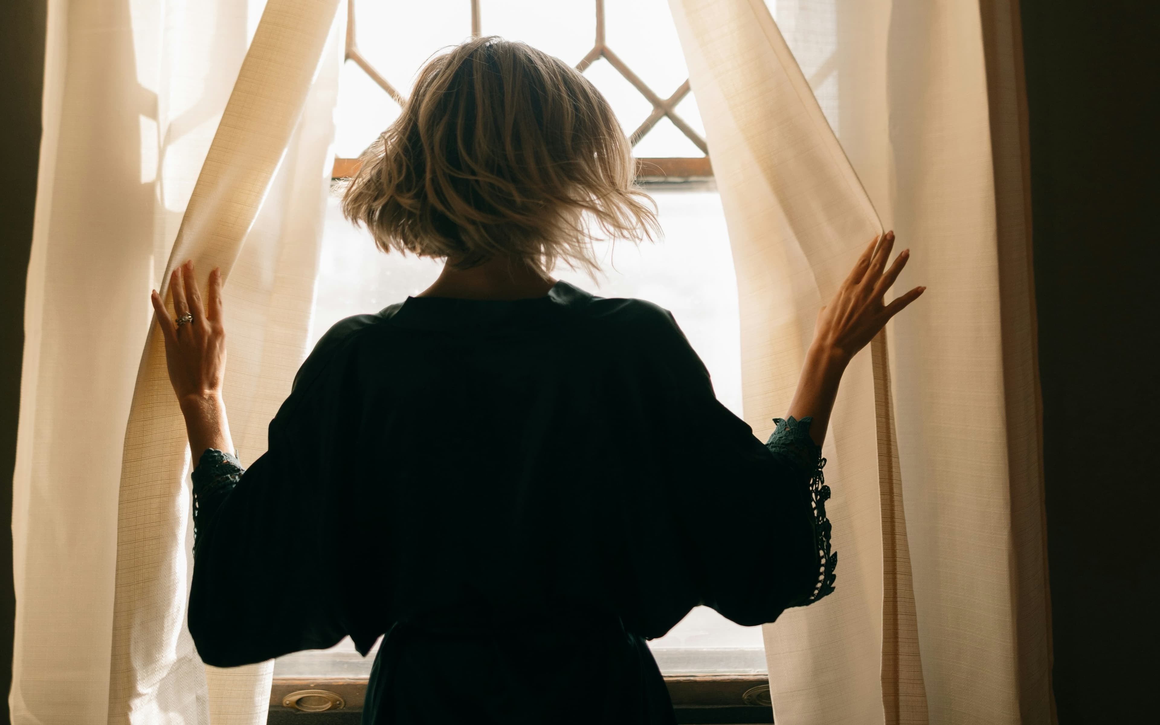 A person with short hair stands by a window, gently holding open sheer curtains, allowing soft light to illuminate the room.