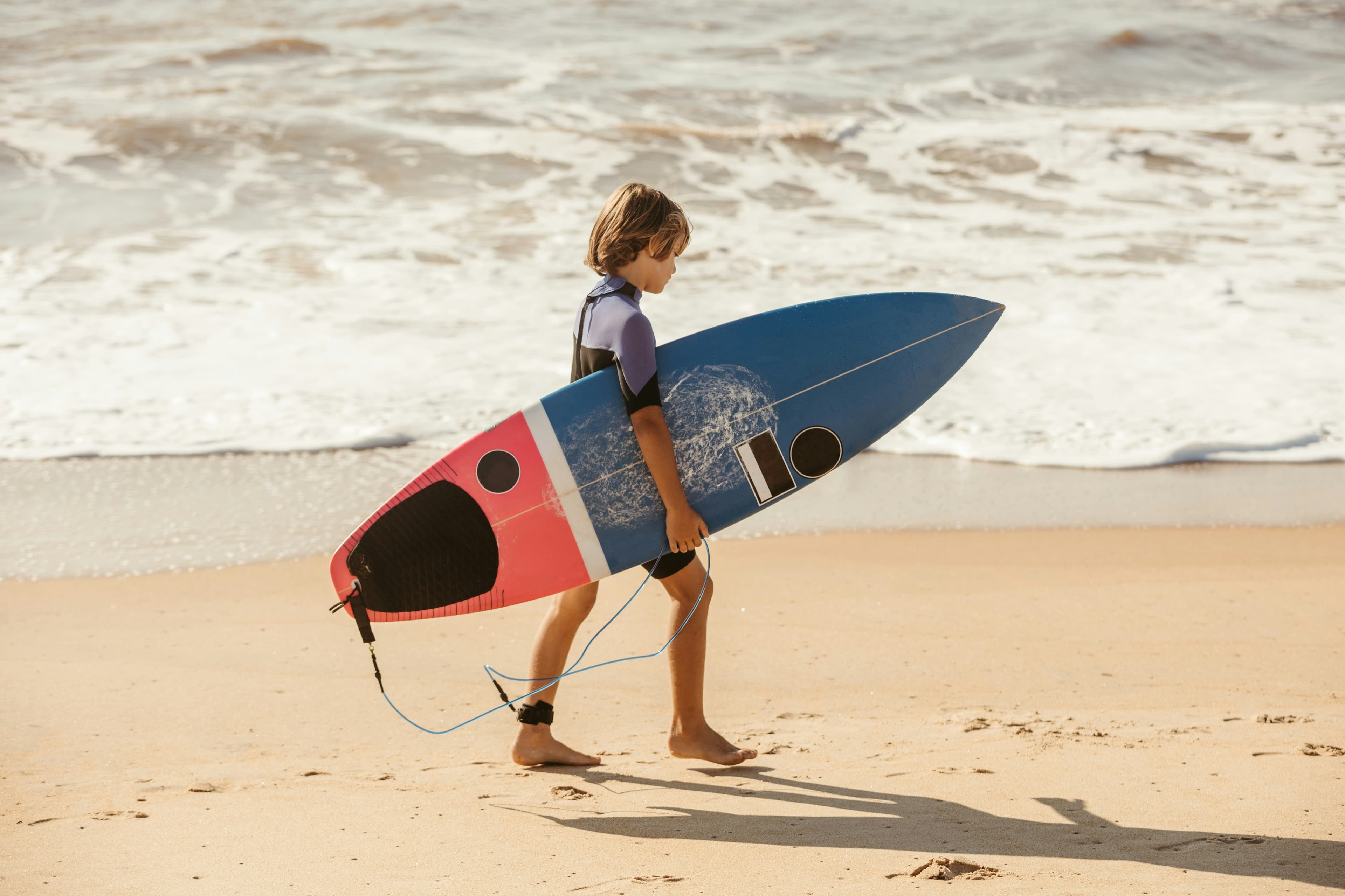 Child walking on a sandy beach carrying a colorful surfboard, with waves in the background.