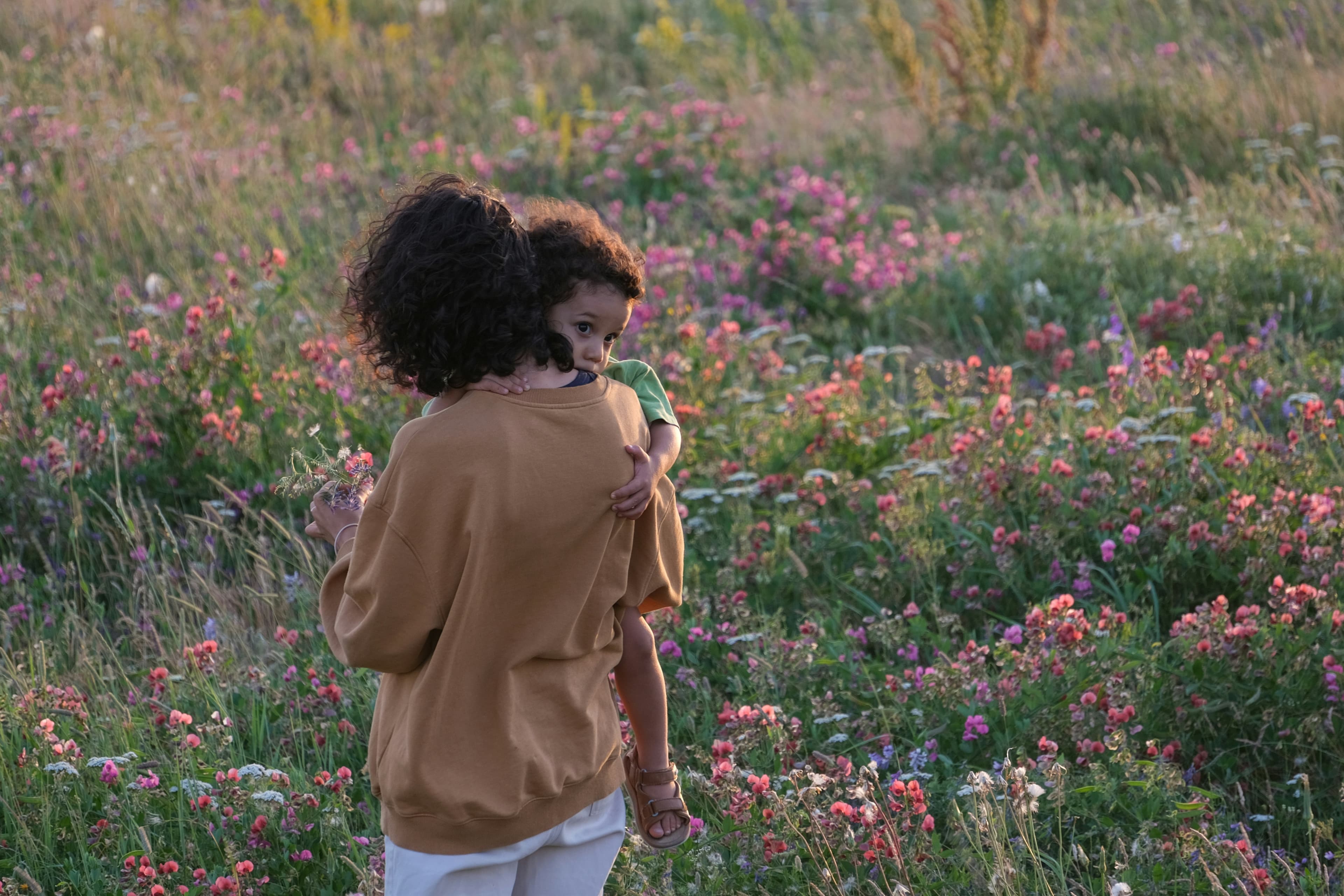 A person with curly hair holds a child in a field of colorful wildflowers under a soft, natural light.